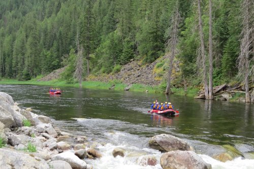 Two rafts with people navigate a calm river surrounded by lush green trees and rocky banks.