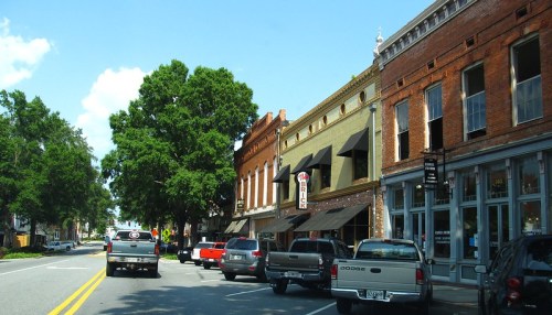 A street view of a small town with shops, trees, and parked trucks under a clear blue sky.