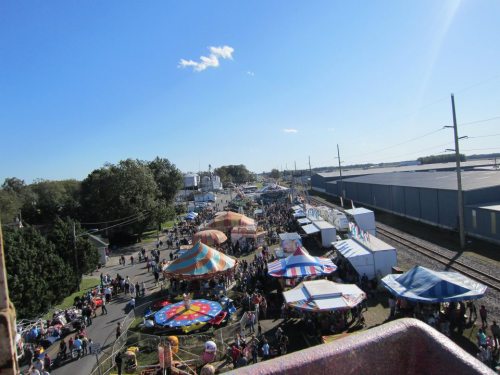 Aerial view of a bustling fair with colorful tents, rides, and crowds under a clear blue sky.