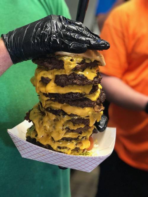 A towering stack of cheeseburgers held by a person wearing a black glove, with melted cheese dripping down.