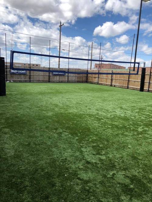 A green turf sports field with a volleyball net, surrounded by a wooden fence and a blue sky with clouds.