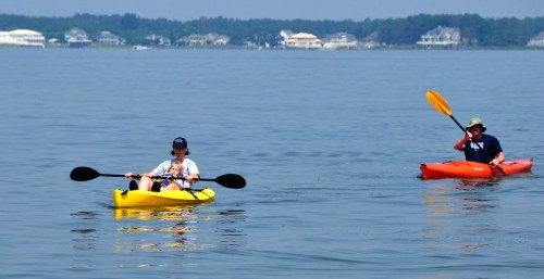 Two people kayaking on a calm lake, one in a yellow kayak with a child, the other in an orange kayak.