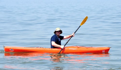 A person in a hat paddles an orange kayak on calm water, enjoying a sunny day outdoors.