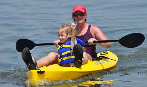 A woman and a girl in a yellow kayak, both smiling, enjoying a sunny day on the water. The girl is playfully splashing.