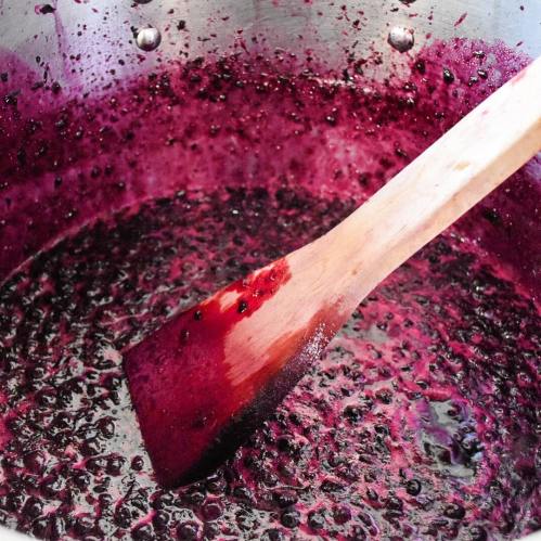 A wooden spoon rests in a pot filled with dark purple berry mixture, showing a rich, thick texture.