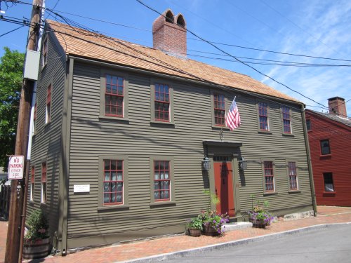 Historic green house with red windows and door, featuring an American flag, surrounded by flowers and brick pathway.