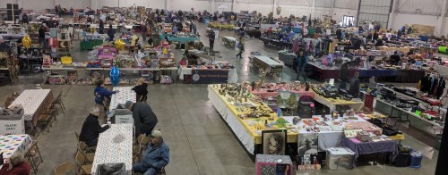 A spacious indoor market filled with vendor tables showcasing various items, with shoppers browsing throughout.