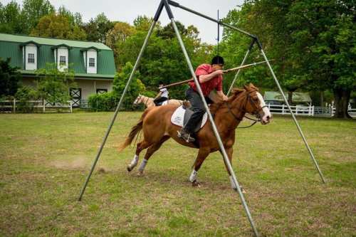 A rider on a horse practices skills under a large frame in a grassy area with a barn in the background.