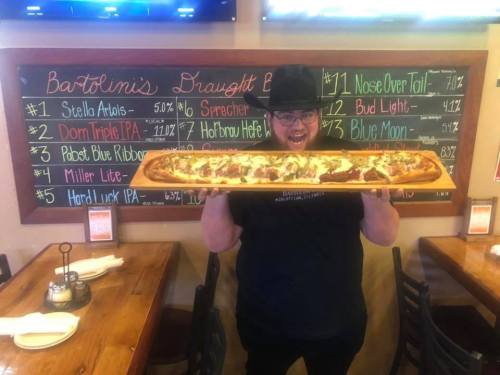 A person wearing a hat holds a large pizza on a wooden board in a restaurant with a colorful menu in the background.
