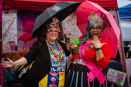 Two women in colorful outfits and accessories, holding umbrellas, smiling and posing at a festive outdoor event.