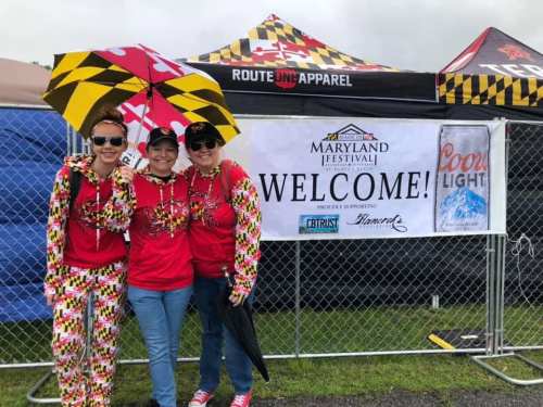 Three women in colorful outfits stand under an umbrella, smiling in front of a "Welcome" banner at the Maryland Festival.