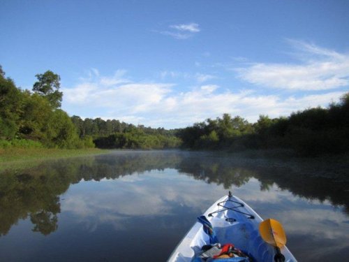 A kayak on a calm river, surrounded by lush greenery and a clear blue sky, with mist rising from the water.