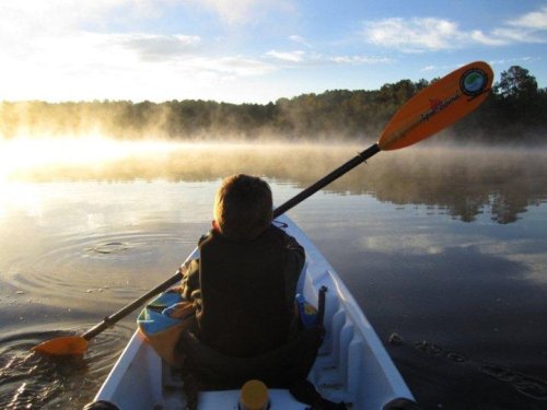 A child paddles a kayak on a misty lake at sunrise, surrounded by trees and calm water.