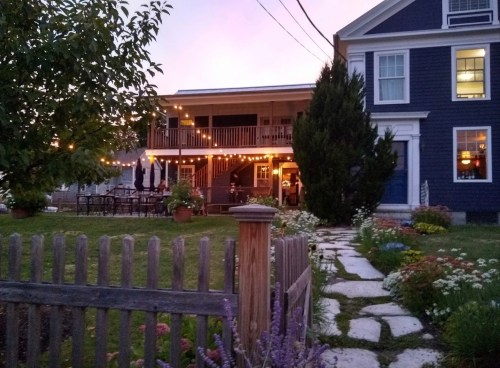 A charming house with a porch, string lights, and a garden, set against a twilight sky.