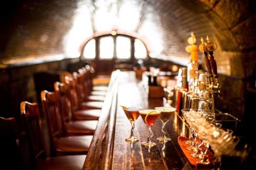 A dimly lit bar with a wooden counter, featuring various cocktails and a row of chairs in a cozy, arched setting.