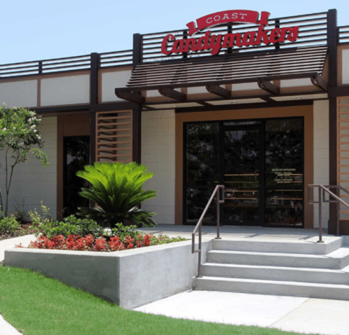 Exterior of Coast Candymakers shop, featuring a sign, steps, and landscaped area with plants and flowers.