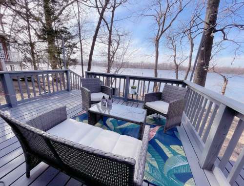 Cozy outdoor seating area with wicker chairs and a table, overlooking a serene lake surrounded by trees.