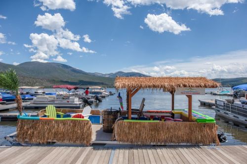 A colorful dock with a thatched-roof bar, surrounded by boats and mountains under a blue sky with fluffy clouds.