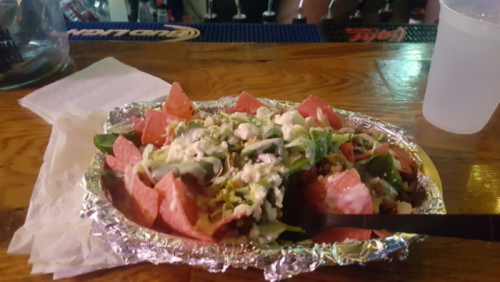 A plate of nachos topped with cheese, lettuce, and jalapeños, served on a foil tray at a bar.