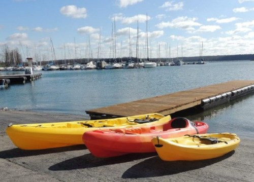 Three colorful kayaks (yellow and red) are parked on a dock by a calm marina with sailboats in the background.