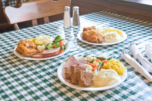 Three plates of comfort food on a checkered tablecloth, featuring fried chicken, ham, vegetables, and mashed potatoes.