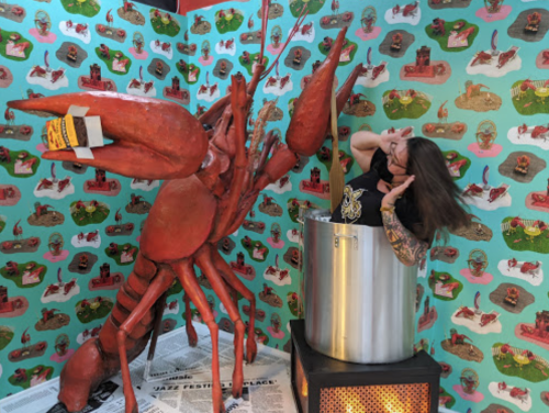 A person with long hair reacts playfully next to a large lobster sculpture beside a metallic pot in a colorful, patterned room.