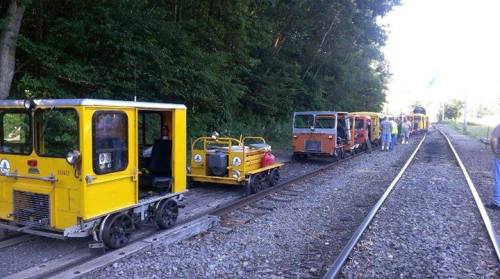 A line of colorful rail cars parked on a gravel track, surrounded by trees and people working nearby.