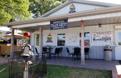 Exterior of a train depot restaurant with a sign, outdoor seating, and a bell in front.