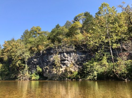 A serene river scene with a rocky cliff and lush trees under a clear blue sky.