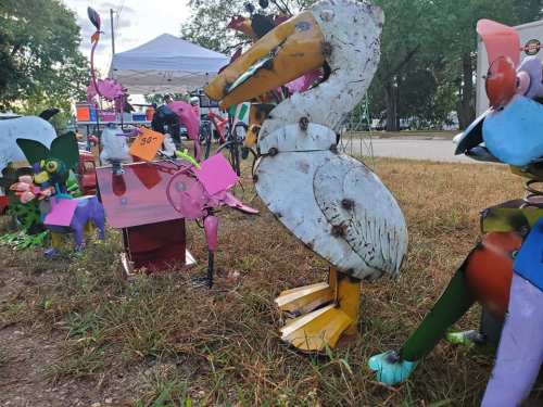 Colorful metal sculptures of birds and animals displayed on grass, with a tent in the background.