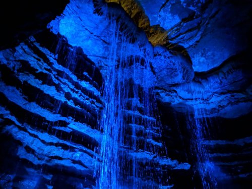 A blue-lit cave with cascading water and layered rock formations, creating a mystical underground scene.