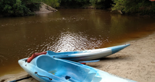 Two blue kayaks on a sandy riverbank, with calm brown water and lush greenery in the background.
