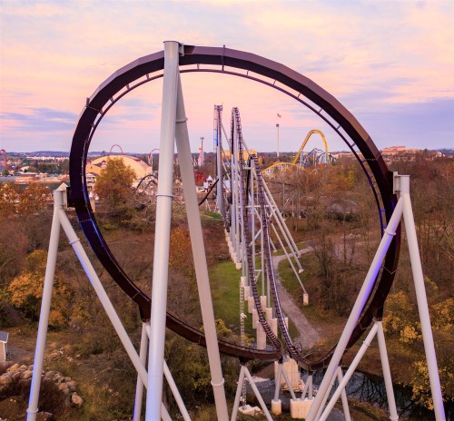 Aerial view of a roller coaster loop with tracks winding through a park, surrounded by trees and a colorful sky.