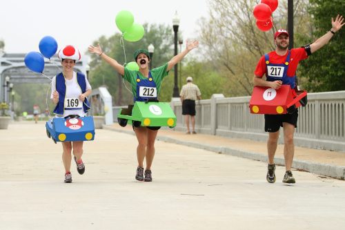 Three runners in colorful Mario Kart costumes joyfully race with balloons on a path, smiling and waving.