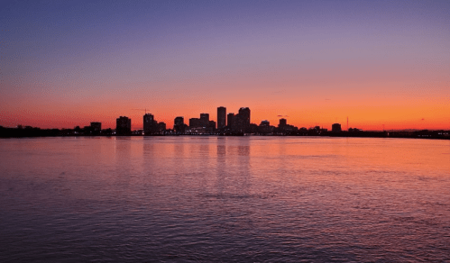 Silhouette of a city skyline at sunset, with vibrant orange and purple hues reflecting on the water.
