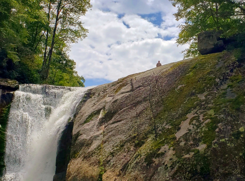 A person sits atop a rocky ledge overlooking a waterfall surrounded by lush greenery and a cloudy sky.