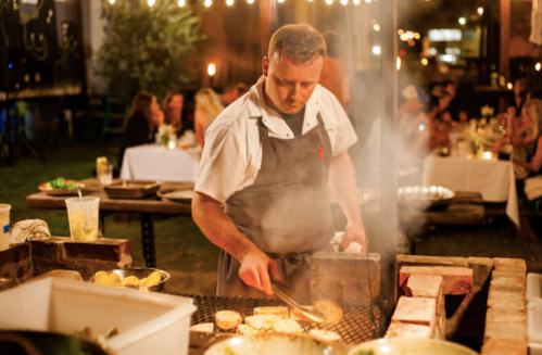 A chef grills food outdoors at a lively evening event, with guests and string lights in the background.