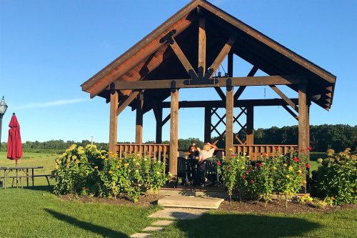 A wooden gazebo surrounded by flowers, with people sitting at a table enjoying the outdoors on a sunny day.