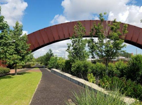 A pathway lined with greenery leads under a large, curved metal arch against a blue sky with fluffy clouds.