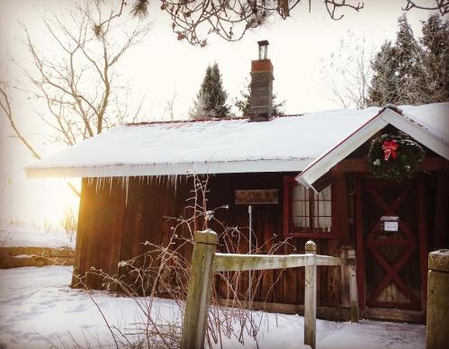 A cozy wooden cabin covered in snow, with icicles hanging from the roof and a festive wreath on the door.
