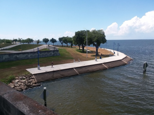 A scenic waterfront path with steps leading to the water, lined by trees and blue lampposts under a clear sky.