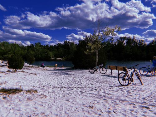 A sandy area by a calm lake, with trees, clouds, bicycles, and people enjoying the outdoors.