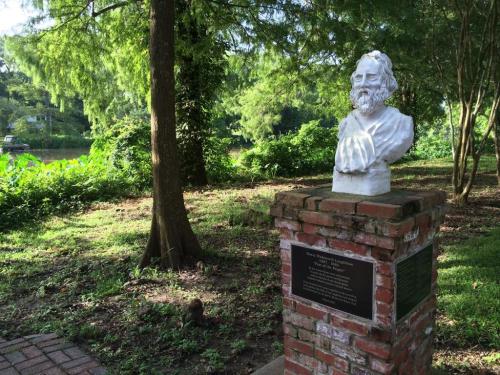 A white bust of a man on a brick pedestal, surrounded by greenery and trees near a body of water.