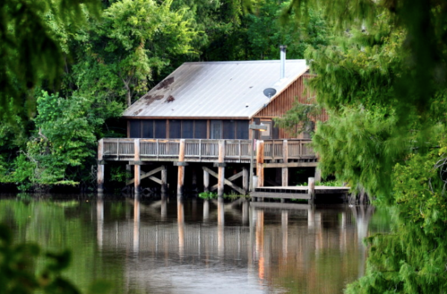 A wooden cabin on stilts by a calm river, surrounded by lush green trees and reflecting in the water.