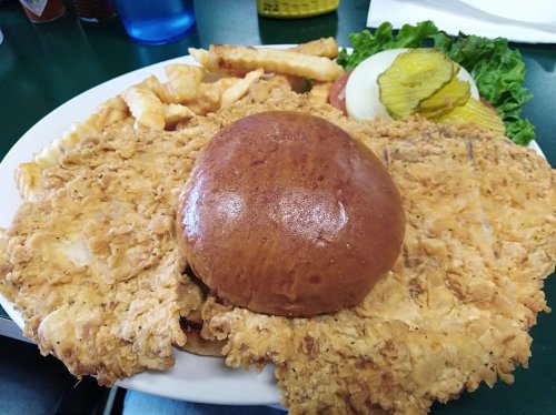 A large fried chicken sandwich on a bun, served with fries, lettuce, tomato, and pickles on a white plate.