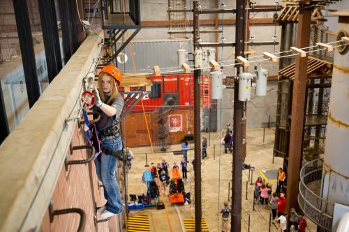 A girl in an orange helmet climbs a vertical wall indoors, with people and equipment visible below.
