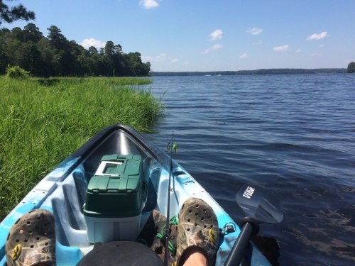 A person’s feet in crocs rest on a kayak, with a cooler and fishing rod, overlooking a calm lake and green shoreline.