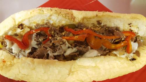 A close-up of a loaded sandwich with sliced beef, peppers, and melted cheese in a toasted hoagie roll on a red plate.