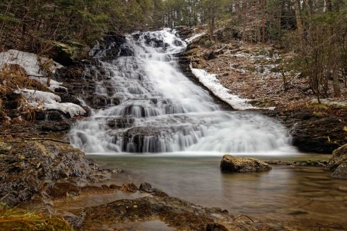 A serene waterfall cascades over rocks into a calm pool, surrounded by trees and remnants of snow.