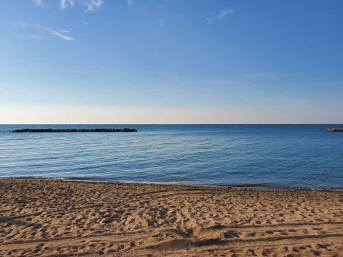 A serene beach scene with calm blue water, a clear sky, and a sandy shore. Rocks are visible in the distance.